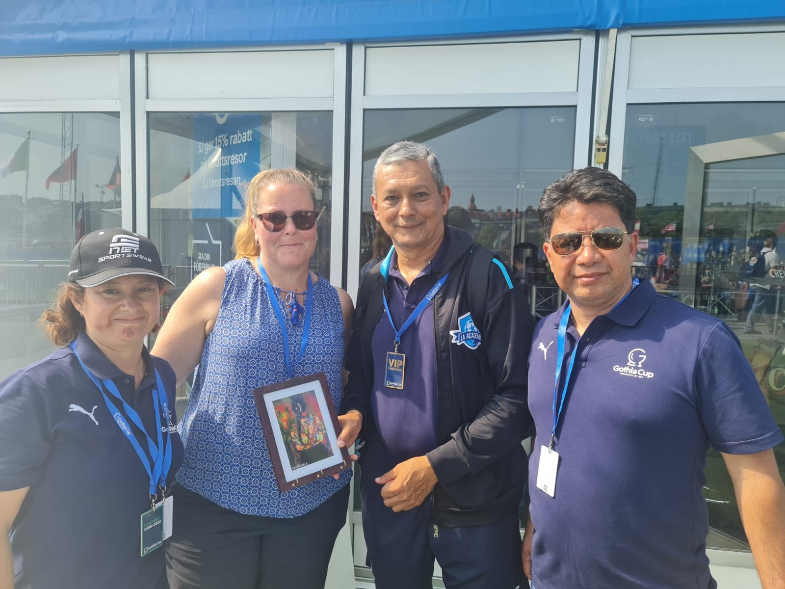 Amigos de Suecia: cuatro personas con credenciales de la Gothia Cup posan juntas; una de ellas sostiene un cuadro de regalo.
